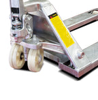 Close-up of a hydraulic pallet jack with silver metal body and yellow warning label; two sturdy white rubber wheels visible.